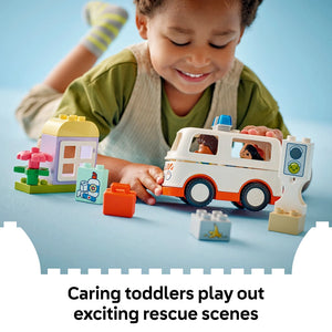 Young child playing with colorful LEGO Duplo ambulance set featuring white and orange ambulance vehicle with blue siren, small figurines, building blocks in green yellow and pink, and accessories on blue surface against light blue background