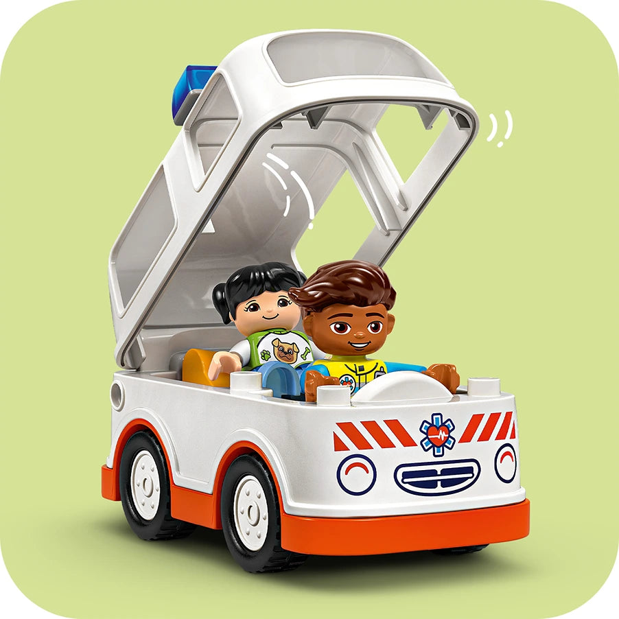 LEGO Duplo ambulance toy set featuring white and red emergency vehicle with opening hood, two smiling child minifigures, orange and brown hair, seated in cabin, displayed against pale green background