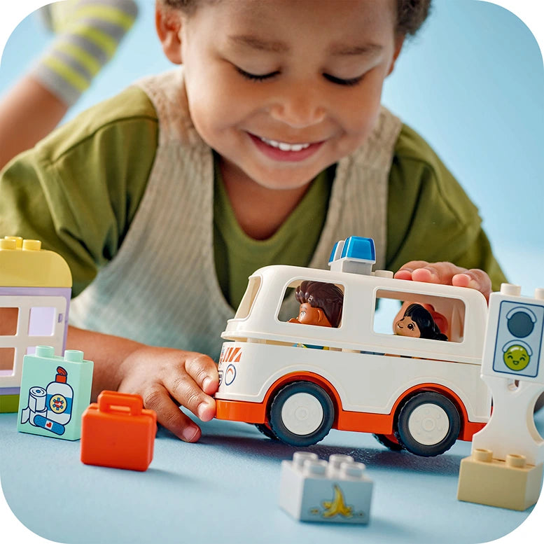 Young child playing with LEGO Duplo white and orange ambulance toy with driver figures, surrounded by colorful building blocks on blue surface