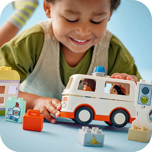 Young child playing with LEGO Duplo white and orange ambulance toy with driver figures, surrounded by colorful building blocks on blue surface