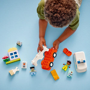 Overhead view of child's hands playing with LEGO Duplo ambulance set 10447, featuring a red and white ambulance with wheels, white ambulance vehicle, minifigures including a medical worker, colorful building blocks in yellow and blue, and various accessories on a light blue background