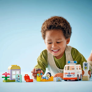 Young child playing with LEGO Duplo ambulance set featuring white and orange ambulance vehicle, figurines, traffic light, and building blocks on blue background