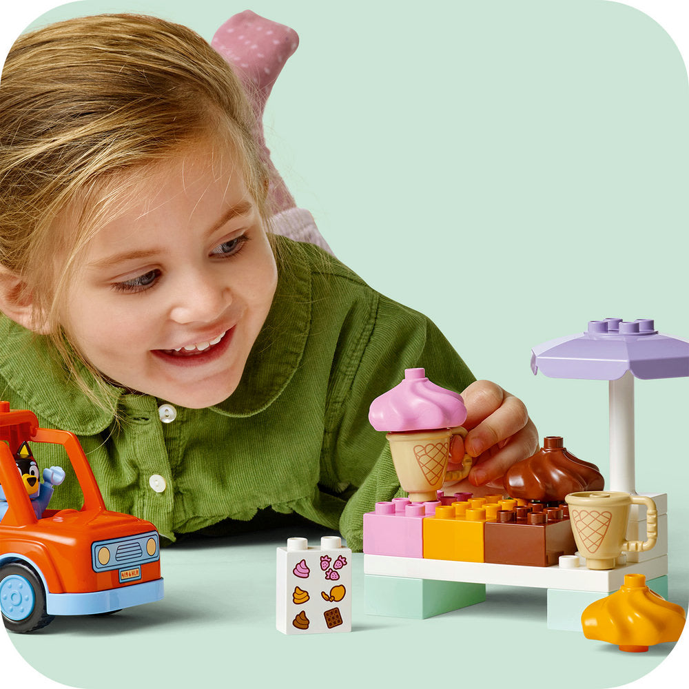 Young child with blonde hair wearing a green cardigan playing with LEGO ice cream shop set featuring pink and white ice cream cones, colorful building blocks in pink yellow and brown, purple parasol, orange toy truck, and food-decorated brick pieces on light green background