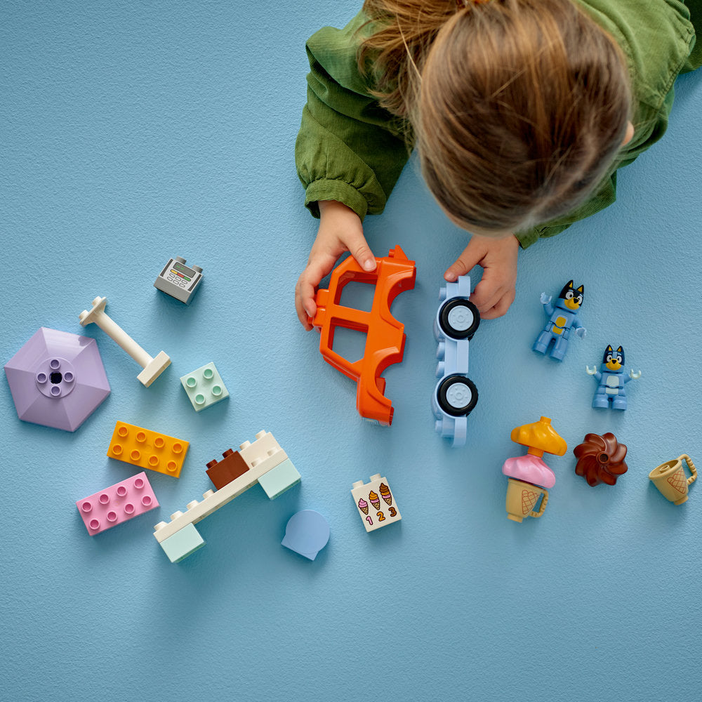 Child in green jacket playing with LEGO set 10458 Ice Cream Trip with Bluey, featuring orange car frame, blue LEGO minifigures, colorful building blocks in pink yellow and cream, ice cream accessories, and various LEGO elements on light blue background