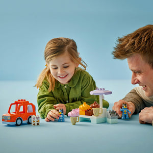 Adult and child playing with LEGO Bluey Ice Cream Trip set 10458 featuring colorful building blocks, red ice cream truck, purple umbrella, and Bluey character minifigures on blue background