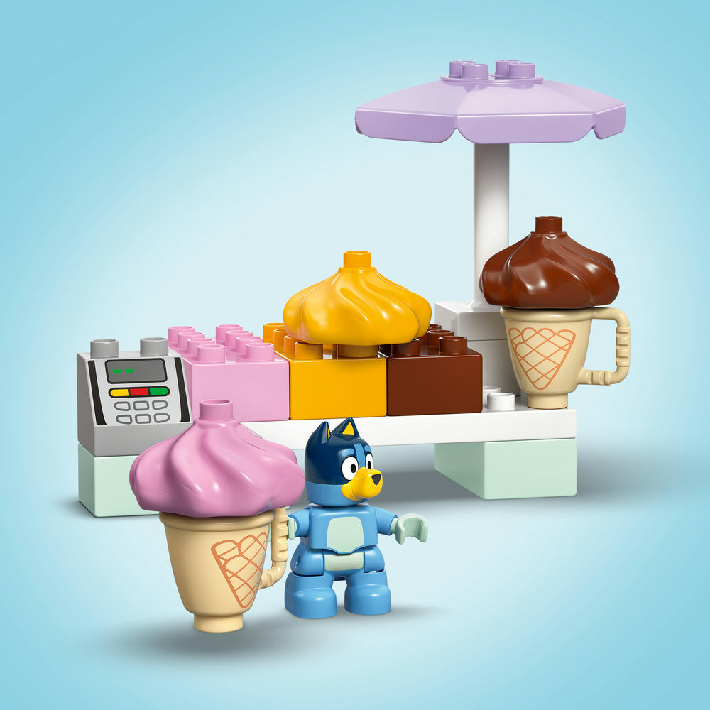 LEGO ice cream shop playset featuring blue Bluey character minifigure, pink and yellow ice cream cones with swirled toppings, brown chocolate ice cream in beige cone-shaped cup, purple umbrella, cash register, and colorful building blocks in pink, yellow and brown on light blue background