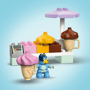LEGO ice cream shop playset featuring blue Bluey character minifigure, pink and yellow ice cream cones with swirled toppings, brown chocolate ice cream in beige cone-shaped cup, purple umbrella, cash register, and colorful building blocks in pink, yellow and brown on light blue background