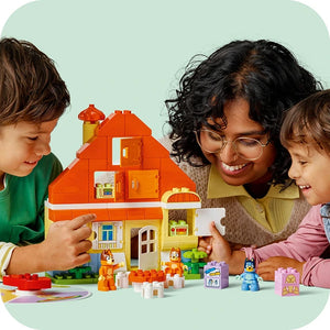 Three children playing with LEGO Bluey's Family House set, a colorful orange and yellow brick building with multiple rooms and windows, surrounded by small LEGO figurines and accessories on a light green background