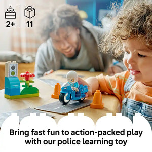 Young child in orange shirt playing with blue LEGO police motorcycle set at wooden table with elderly caregiver, featuring traffic cones and flower elements, age 2+ with 11 pieces