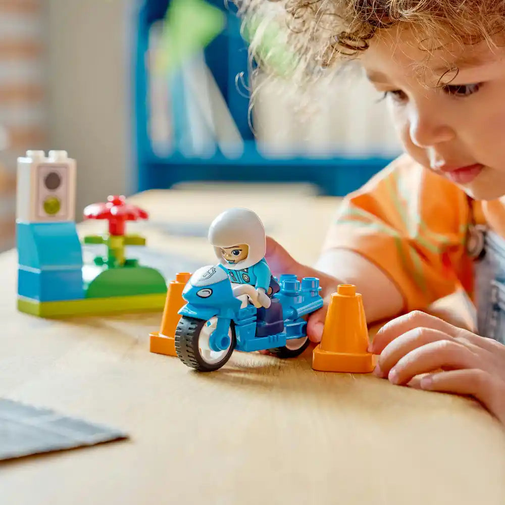 Young child with curly hair playing with LEGO blue police motorcycle set featuring a minifigure officer, blue motorcycle with white wheels, orange traffic cones, and colorful building blocks on wooden table