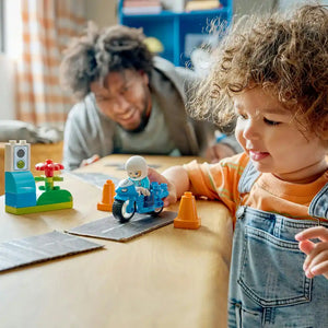 Young child with curly blonde hair playing with LEGO blue police motorcycle set featuring minifigure, orange traffic cones and building blocks on wooden table with caregiver in background