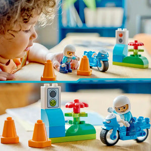 Young child with curly blonde hair playing with LEGO Duplo blue police motorcycle set featuring white police officer minifigure, blue motorcycle, orange traffic cones, traffic light, and green flower piece on wooden surface with blue storage bin in background