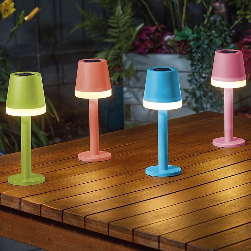 Four solar-powered table lamps in assorted colors - lime green, coral orange, sky blue, and pink - with illuminated white bases arranged on a wooden deck table with potted plants and greenery in the background