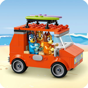 LEGO Bluey's Beach & Family Car Trip building set featuring a bright orange brick vehicle with black wheels on sandy beach background, with cartoon dog characters visible in the cabin and a green and purple surfboard on the roof rack against a blue sky and ocean backdrop