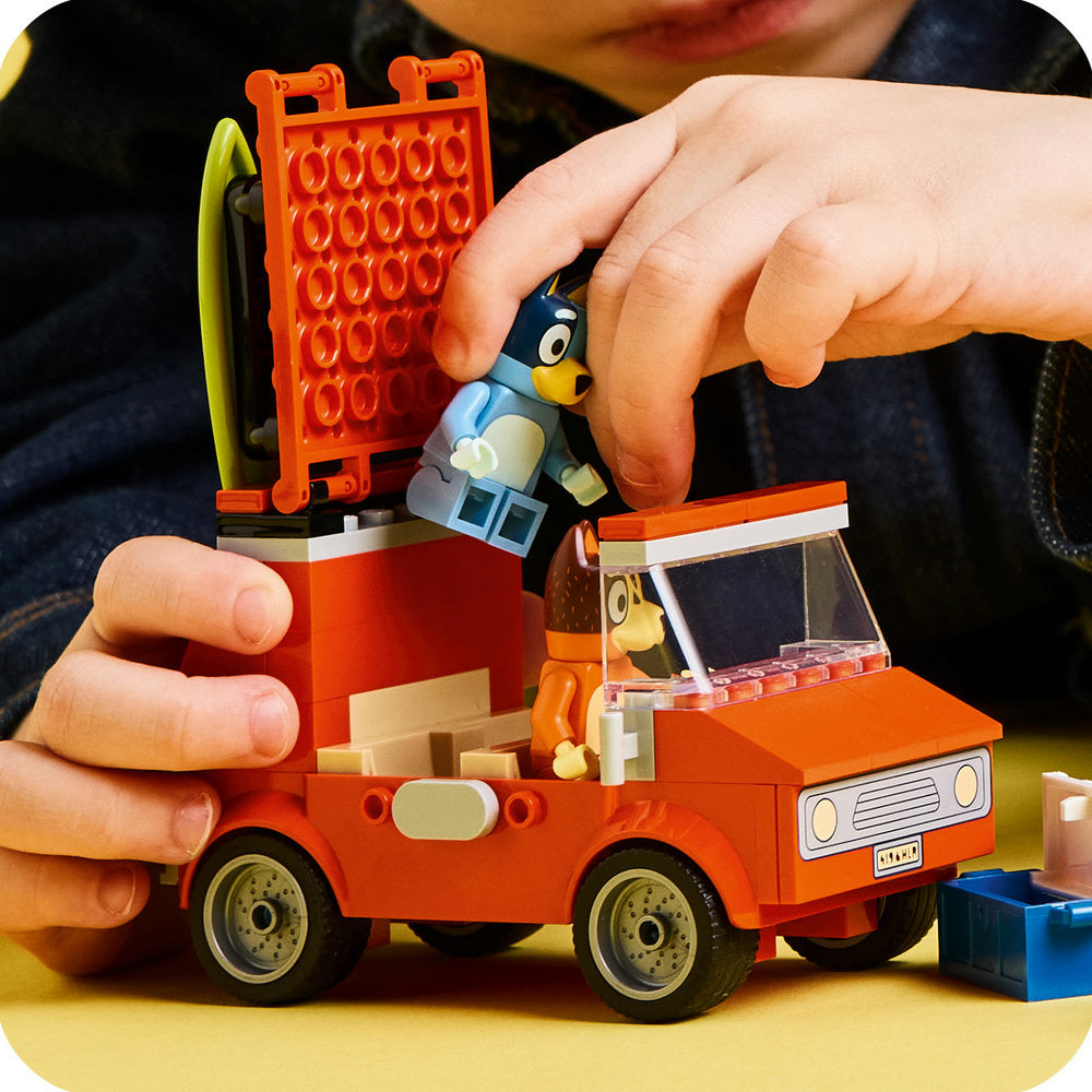 Child's hands assembling a LEGO set featuring a bright orange fire truck with yellow wheels and a matching orange ladder structure, with a small blue bird character figure visible in the play scene on a yellow surface