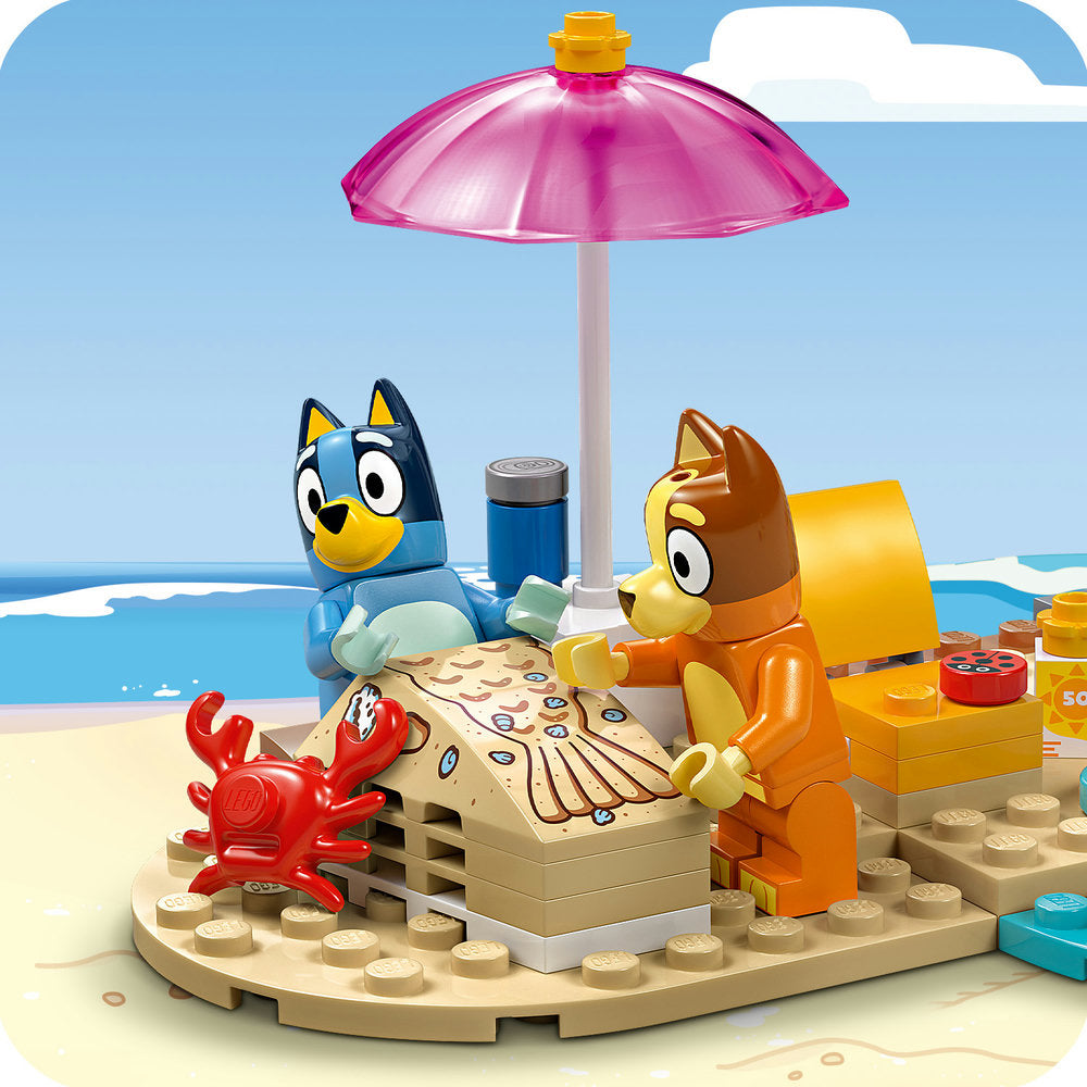 LEGO Bluey beach playset featuring blue Bluey and orange Bingo minifigures under a pink umbrella with a red crab, picnic basket, beverage container, and sand-colored baseplate against a blue ocean and sky background