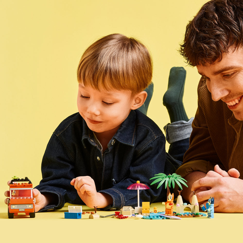 Adult and young child playing together with colorful LEGO Bluey beach and family car trip building set on yellow surface, featuring miniature figures, umbrellas, vehicle pieces and accessories