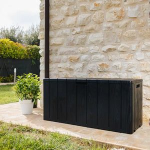 Black wooden garden storage box positioned on a stone patio next to a cream stone wall, with a potted green plant and manicured hedges visible in the background