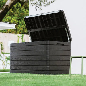 Toomax Dolomiti anthracite garden storage box with open lid showing black interior compartments, positioned on grass in a garden setting with white fencing and green foliage in background