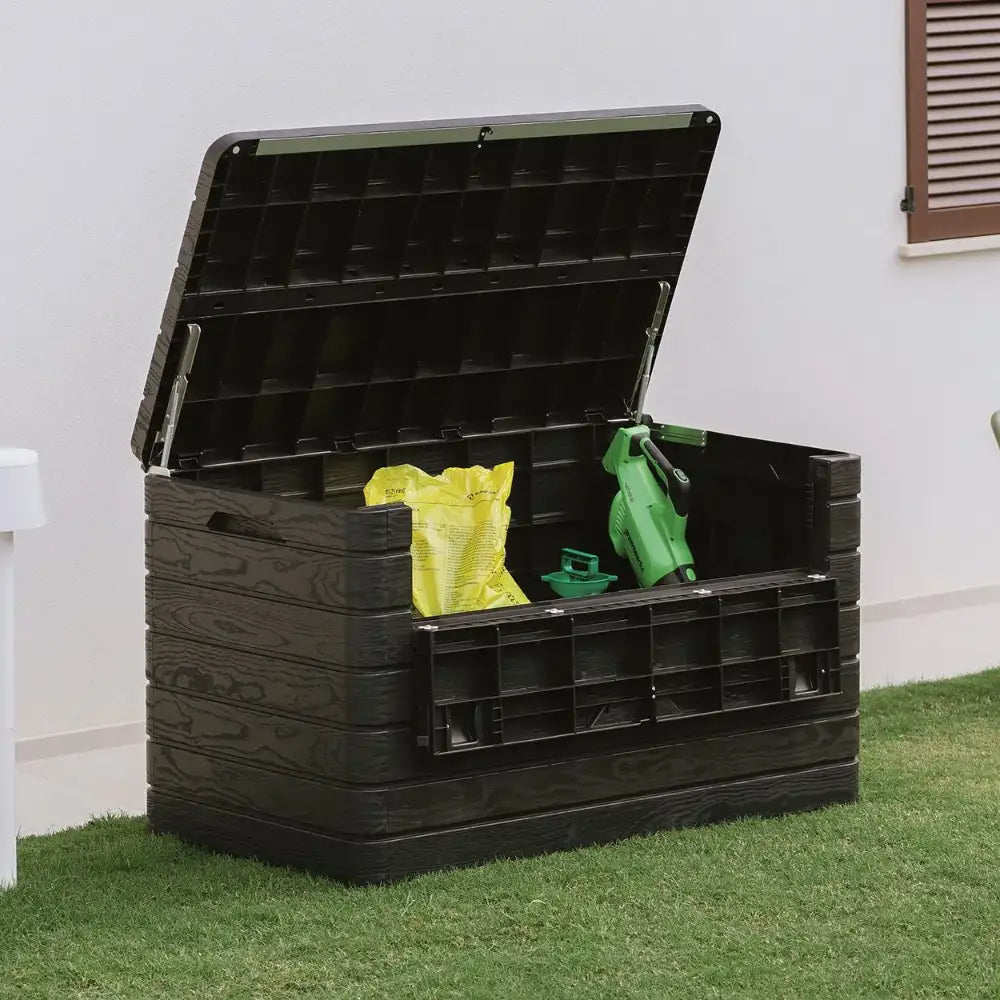 Black plastic garden storage box with open lid containing yellow bags and green garden tools, positioned on grass against white house wall with brown window shutter