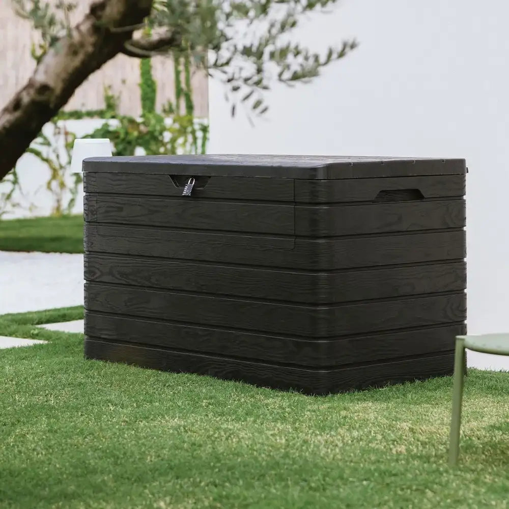 Black resin garden storage box with horizontal ribbed design sitting on grass with tree and white fence in background