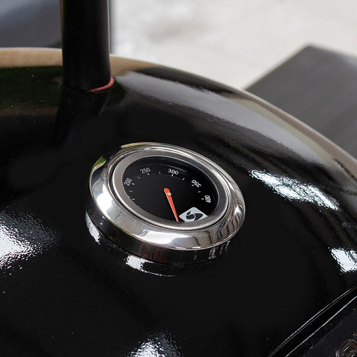 Close-up of a chrome temperature gauge mounted on a black glossy charcoal barbecue, displaying a scale from 200 to 400 degrees Fahrenheit with a red indicator needle pointing to approximately 300 degrees