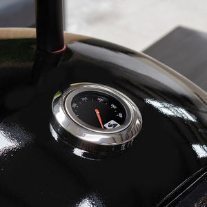 Close-up of a chrome temperature gauge mounted on a black glossy charcoal barbecue, displaying a scale from 200 to 400 degrees Fahrenheit with a red indicator needle pointing to approximately 300 degrees