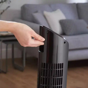 Hand operating the touch control panel on a black Dimplex Mont Blanc tower fan in a living room setting with gray sofa and wooden floor