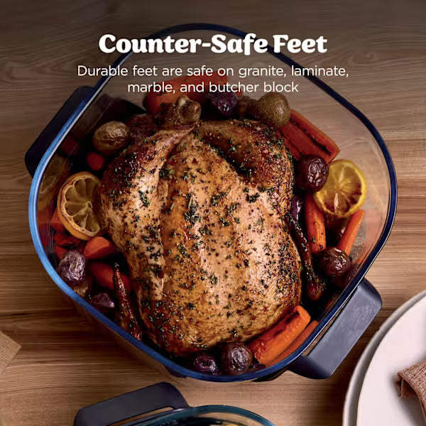 Overhead view of a roasted chicken with herbs and lemon slices in a blue-rimmed glass Ninja air fryer basket surrounded by roasted vegetables, demonstrating counter-safe feet on a wooden surface