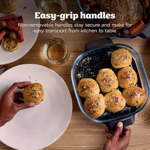 Overhead view of Ninja CRISPi PRO XL Glass Air Fryer with blue handles displaying golden cooked rolls, demonstrating easy-grip handles for transport from kitchen to table, with white plate and serving accessories on wooden surface