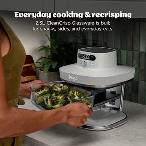 Ninja CRISPi PRO XL Glass Air Fryer in white and bone with digital display showing 2000 temperature setting, displaying roasted broccoli in 2.3L glassware bowl on kitchen counter with person in olive green top holding tray