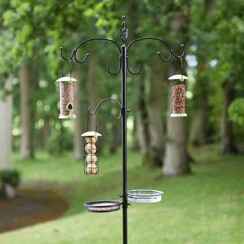 Black metal bird feeding station with multiple hanging feeders in garden setting, featuring two seed feeders and one suet feeder suspended from decorative scrolled arms, with two circular trays at base, surrounded by blurred green trees and lawn