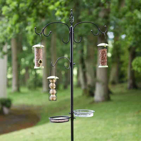 Black metal bird feeding station with multiple hanging feeders in garden setting, featuring two seed feeders and one suet feeder suspended from decorative scrolled arms, with two circular trays at base, surrounded by blurred green trees and lawn