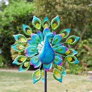 Colorful metal peacock solar wind spinner with turquoise blue body, green and yellow feather details, and purple accents on black pole in garden setting