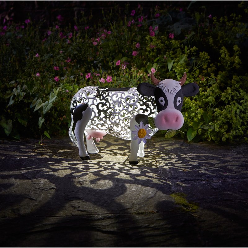 Smart Solar Garden Silhouette Daisy Cow outdoor light featuring white and black decorative metal cow with pink nose and horns, illuminated at night in a garden setting surrounded by pink and purple flowers and green foliage
