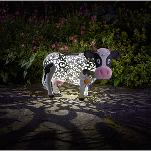 Smart Solar Garden Silhouette Daisy Cow outdoor light featuring white and black decorative metal cow with pink nose and horns, illuminated at night in a garden setting surrounded by pink and purple flowers and green foliage