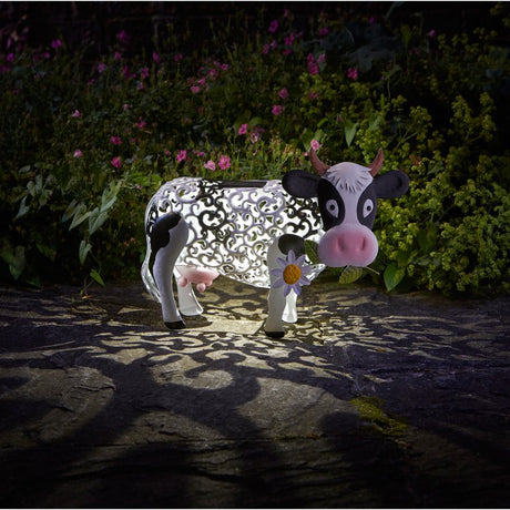 Smart Solar Garden Silhouette Daisy Cow outdoor light featuring white and black decorative metal cow with pink nose and horns, illuminated at night in a garden setting surrounded by pink and purple flowers and green foliage