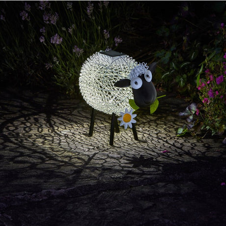 Solar-powered decorative sheep garden light with illuminated white woolly body, black head and legs, blue eyes, and yellow daisy flower detail, displayed at night among flowering plants and lavender in a garden setting