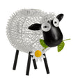 Solar-powered decorative garden sheep silhouette with white textured woolly body, black head and ears, large googly eyes, green leaves in mouth, white daisy flower on chest with yellow center, and four black metal legs