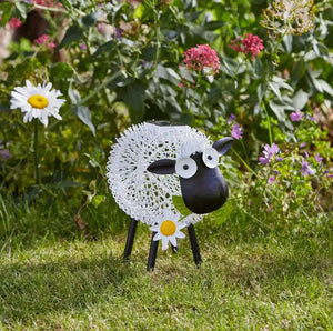 Decorative garden sheep silhouette with white lace wool body, black head and legs, large googly eyes, and white daisy flower detail, positioned in grass with colorful garden flowers in background