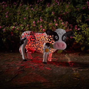 Decorative garden silhouette of a cow with red and orange patterned body, white legs and face, pink nose, and white horns, holding a white daisy flower, positioned in front of pink flowering plants