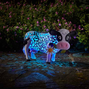 A glowing neon blue cow garden ornament with cyan paisley pattern and white face details, standing on dark ground with purple flowers blooming in the background, illuminated by solar-powered LED lights