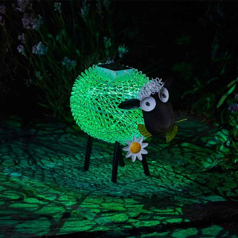 Illuminated solar garden sheep ornament with bright green glowing fleece, black body and legs, white daisy flower detail, and moving googly eyes, positioned on dark ground with plants in background at night