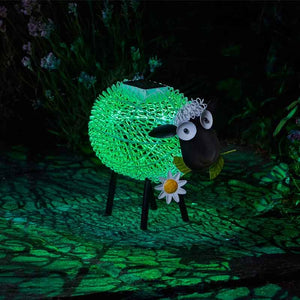 Illuminated solar garden sheep ornament with bright green glowing fleece, black body and legs, white daisy flower detail, and moving googly eyes, positioned on dark ground with plants in background at night