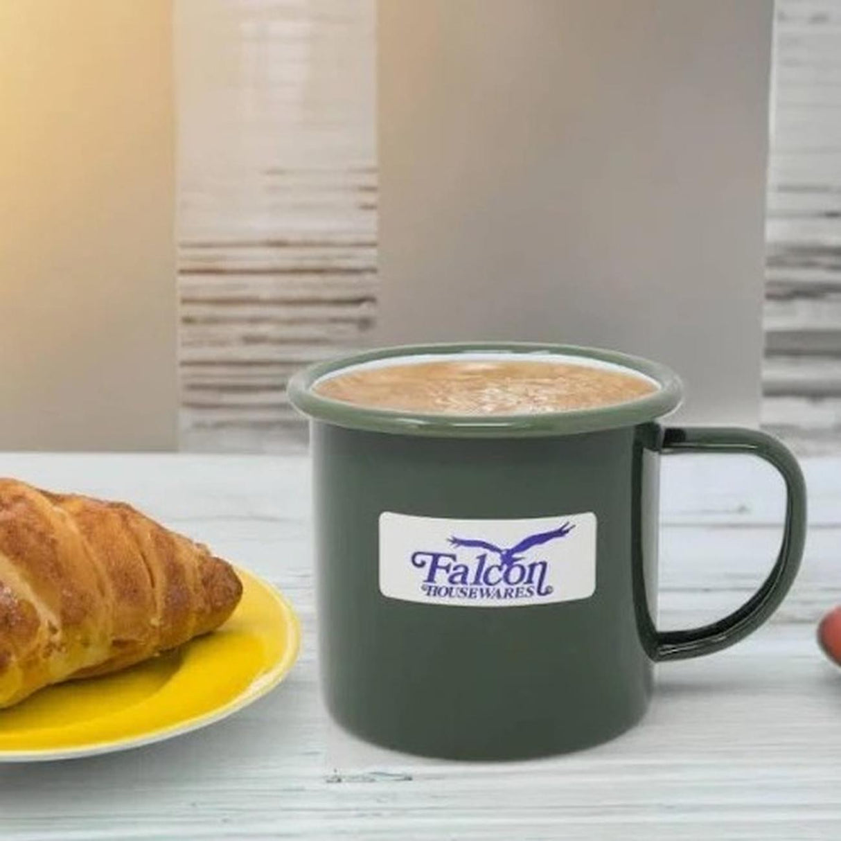 Falcon green enamel mug with white logo containing hot beverage, displayed on white desk next to croissant on yellow plate and open documents in background