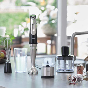 Braun MultiQuick 9 hand blender with stainless steel shaft and black top displayed on kitchen counter with five attachments including whisk, chopper bowl, and measuring cup, surrounded by fresh ingredients and flowers in bright window setting