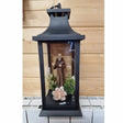 Black metal lantern with Padre Pio figurine, white flowers and green foliage inside, wooden background, 27cm tall religious memorial ornament