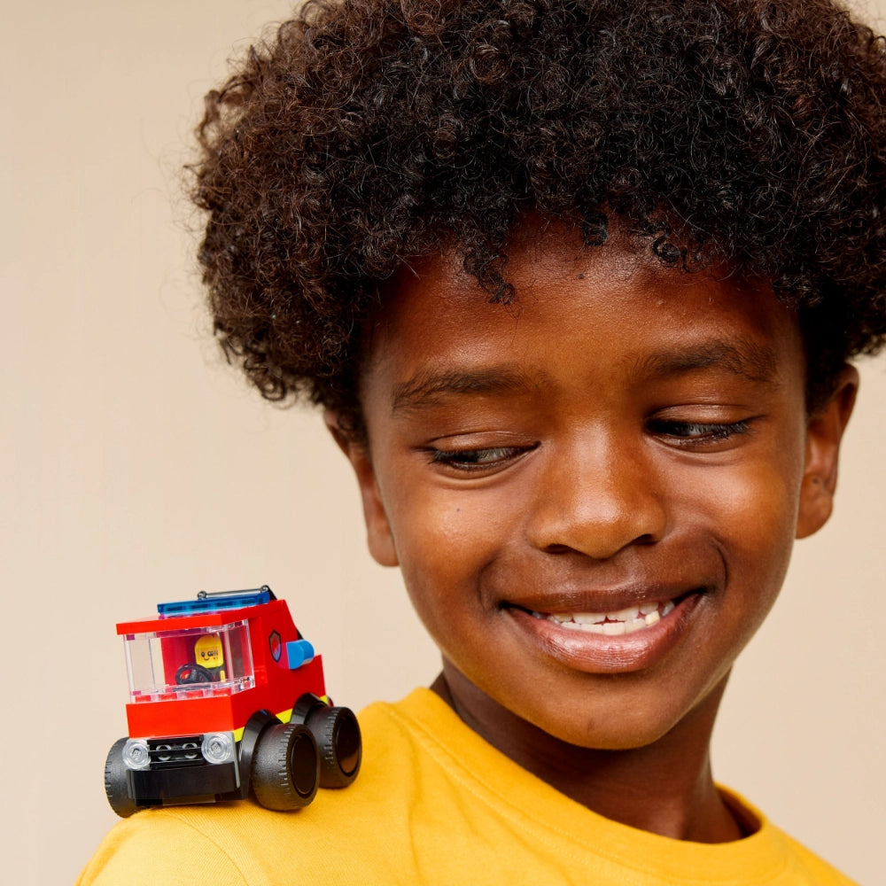 Young child wearing yellow shirt smiling while holding red and blue LEGO fire truck toy with black wheels against neutral beige background