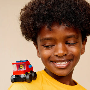 Young child wearing yellow shirt smiling while holding red and blue LEGO fire truck toy with black wheels against neutral beige background
