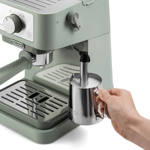 DeLonghi Stilosa EC260.GR espresso machine in sage green with stainless steel milk frother pitcher being held by hand, featuring control knobs and drip tray